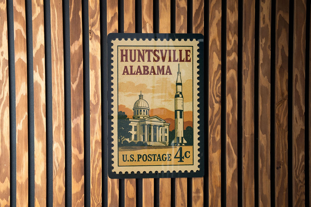A vintage U.S. postage stamp from Huntsville, Alabama, featuring a rocket ship and a building, mounted on a wooden wall.