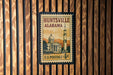 A vintage U.S. postage stamp from Huntsville, Alabama, featuring a rocket ship and a building, mounted on a wooden wall.