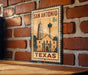 A vintage-style metal sign depicting the San Antonio, Texas skyline, with a building and a tower, mounted on a brick wall.