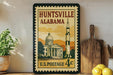 A vintage U.S. postage stamp from Alabama featuring a building and a rocket ship, displayed on a shelf next to a wooden cutting board and a potted plant.
