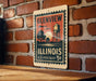 A vintage postage stamp from the United States, featuring a scene of a water tower and trees, mounted on a brick wall.
