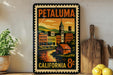 A vintage-style metal sign with a colorful illustration of a cityscape and a clock, mounted on a wall next to a wooden cutting board and some potted plants.