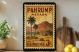 A framed vintage U.S. postage stamp depicting a desert landscape with palm trees and a house, placed on a shelf next to a wooden cutting board and a potted plant.