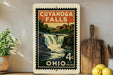 a vintage-style postage stamp from Ohio, depicting a waterfall and a town, placed on a shelf next to a wooden cutting board and some lemons.