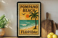 A framed poster of Pompano Beach, Florida, featuring a palm tree, ocean, and sunset, is displayed on a wall next to a wooden cutting board and a potted plant.