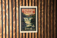 A vintage postage stamp from Ohio featuring a waterfall and a building, mounted on a wooden wall.