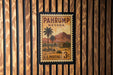 A vintage U.S. postage stamp from Nevada, featuring a desert landscape with palm trees and mountains, mounted on a wooden wall.
