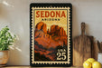 A framed vintage postage stamp depicting Sedona, Arizona, with the text "SEDONA ARIZONA" and "USA 25" printed on it. The stamp is displayed on a shelf next to a wooden cutting board and a potted plant.
