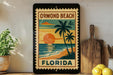 A framed poster of a tropical beach scene with palm trees and a sunset, displayed on a shelf next to a wooden cutting board and some lemons.