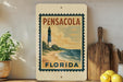 A framed vintage-style poster of a lighthouse in Pensacola, Florida, with the text "PENSACOLA FLORIDA" on it, is displayed on a shelf next to a wooden cutting board and some kitchen utensils.