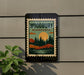 A sign for Woodbury Minnesota, featuring a scenic landscape with a lake, mountains, and trees, mounted on a wall.