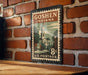 A vintage postage stamp from the United States, featuring an illustration of a building and a clock, mounted on a brick wall.