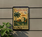 A vintage-style postage stamp from Carolina, featuring a palm tree and buildings, is displayed on a wall next to a plant.