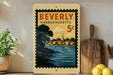A vintage postage stamp from Beverly, Massachusetts, depicting a scenic coastal town with a church steeple, is displayed on a shelf next to a wooden cutting board and a potted plant.