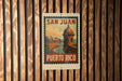 A vintage-style poster of the San Juan, Puerto Rico, featuring a castle-like structure and a body of water, mounted on a wooden wall.