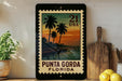 A framed vintage postage stamp depicting a tropical beach scene with palm trees and a sunset, placed on a shelf next to a wooden cutting board and a potted plant.