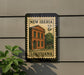 A New Iberia U.S. 6¢ postage stamp from Louisiana, featuring an illustration of a building, is displayed on a wall next to a plant.