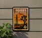 A postage stamp featuring a desert landscape with a tower and a cactus, mounted on a wall.