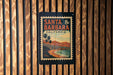 A vintage-style poster of Santa Barbara, California, featuring palm trees and a beach scene, is displayed on a wooden wall.