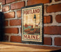 A vintage-style metal stamp depicting a lighthouse and a coastal scene, mounted on a brick wall.
