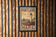A vintage-style postage stamp from Portland, Maine, featuring a lighthouse and a sailboat, is displayed on a wooden wall.