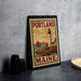 A framed vintage postage stamp from Maine, featuring a lighthouse and sailboat, is displayed on a countertop next to some black camera lenses.