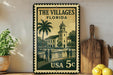 A vintage-style metal sign with a tropical scene depicting a building, palm trees, and people, along with the text "THE VILLAGES FLORIDA" and "USA 5c".
