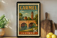 A vintage-style postage stamp from Carmel, Indiana, featuring a bridge and a clock tower, displayed on a shelf with kitchen utensils and a potted plant.
