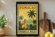 A framed vintage postage stamp depicting a tropical scene with palm trees and a church, hanging on a wall next to a wooden cutting board and a potted plant.