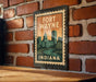 A metal sign with a vintage-style illustration of Fort Wayne, Indiana, mounted on a brick wall.