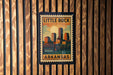 A framed poster of Little Rock, Arkansas, featuring a cityscape and a bridge, is displayed on a wooden wall.