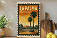 A vintage-style stamp depicting a tropical beach scene with palm trees and a building, placed on a shelf next to a wooden cutting board and a potted plant.