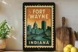 A framed poster of Fort Wayne, Indiana, featuring a cityscape and a clock tower, is displayed on a shelf next to a wooden cutting board and a potted plant.