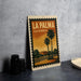 A vintage-style metal sign with the text "LA PALMA CALIFORNIA" and an illustration of a palm tree and a building, placed on a gray countertop next to some black camera lenses.
