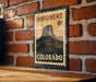 A metal sign with a mountain landscape and the text "MONUMENT 8c COLORADO" is displayed on a brick wall.