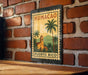 A metal sign with a tropical scene of palm trees and a church is hanging on a brick wall.