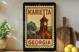 A framed poster of a clock tower in Marietta, Georgia, with the text "MARIETA GEORGIA" on it, displayed on a shelf next to a wooden cutting board and a potted plant.