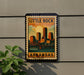 A framed poster of Little Rock, Arkansas, featuring a cityscape with a bridge and a sunset in the background.