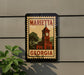 A sign displaying the name "MARIEtta Georgia" with a clock tower illustration, mounted on a wall next to a plant.