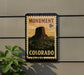 A vintage-style postage stamp depicting the Monument to the Revolution in Colorado, mounted on a wall next to a plant.