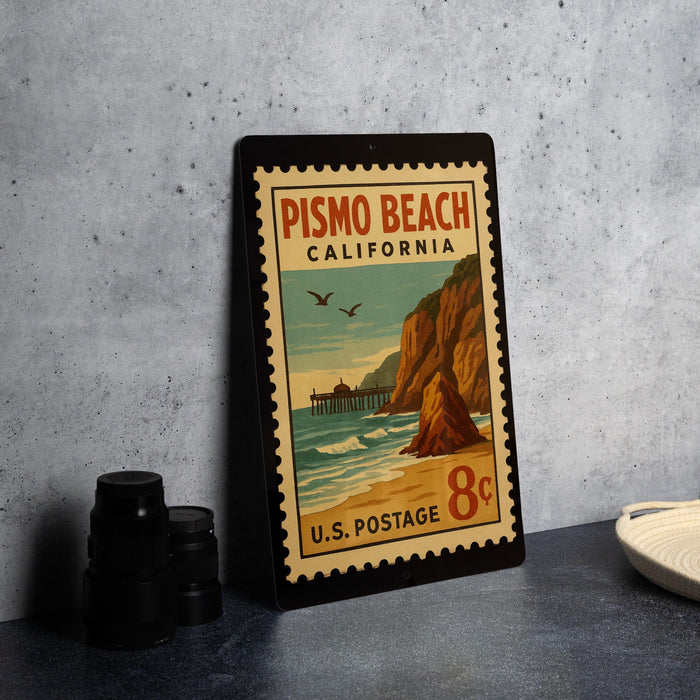 A vintage postage stamp from Pismo Beach, California, featuring a beach scene with a pier and cliffs, placed on a table next to some camera equipment.