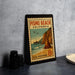 A vintage postage stamp from Pismo Beach, California, featuring a beach scene with a pier and cliffs, placed on a table next to some camera equipment.