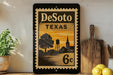 A vintage-style metal stamp with the text "DeSoto, TX 6c" displayed on it, along with a silhouette of a town and a water tower. The stamp is placed on a shelf next to a wooden cutting board and some potted plants.