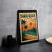 A framed poster of a tropical beach scene with palm trees, a pier, and a sunset is displayed on a table.