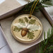 A decorative ornament in a box, featuring a brown acorn surrounded by green leaves, with a pine branch and greenery in the background.