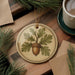 a round ornament with a tree design, including an acorn, hanging on a wooden surface. There is also a cup of coffee and some pine branches in the background.