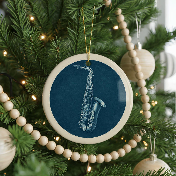 A blue ceramic ornament with a white saxophone design hangs from a Christmas tree, surrounded by white beads and greenery.