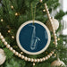 A blue ceramic ornament with a white saxophone design hangs from a Christmas tree, surrounded by white beads and greenery.
