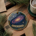 A circular ornament with a galaxy design and the word "ANDROMEDA" on it, placed on a wooden surface next to a cup of coffee and pine branches.