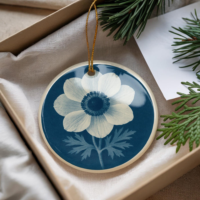 A blue and white ceramic ornament with a white flower design is displayed in a box, accompanied by pine branches and a white card.